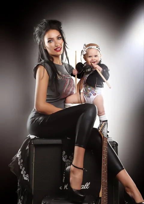 Mother in a rock tee and leather leggings sitting on an amp with her baby in a mini leather jacket holding drumsticks